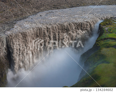 Dettifoss waterfall in Iceland cascades with immense force, creating mist that contrasts with rugged rocks and vibrant green moss covered cliffs. Dettifoss waterfall in Iceland cascades with immense force, creating mist that contrasts with rugged rocks and vibrant green moss covered cliffs. 134284602
