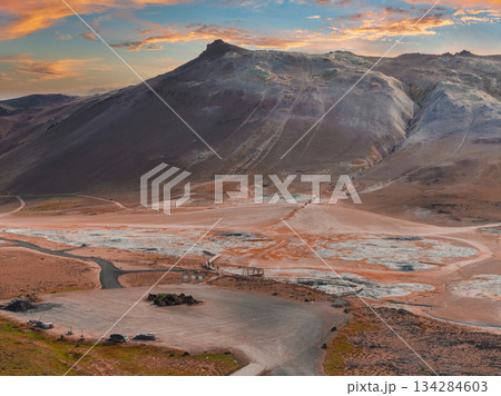 Hverir geothermal area near Lake Myvatn with steaming fumaroles, bubbling mud pots, vibrant soil, a mountain backdrop, and a wooden walkway at sunset. 134284603