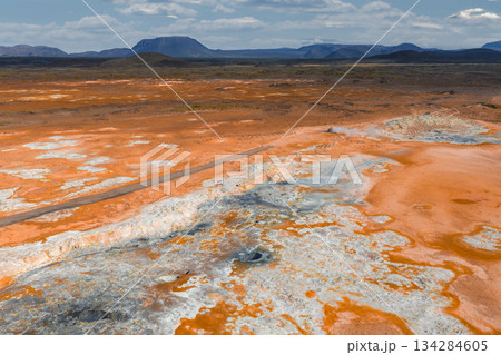 Vibrant geothermal landscape in Iceland with orange and white mineral deposits, steam vents, bubbling mud pots, a road, and rugged mountains under clouds. Vibrant geothermal landscape in Iceland with orange and white mineral deposits, steam vents, bubbling mud pots, a road, and rugged mountains under clouds. 134284605