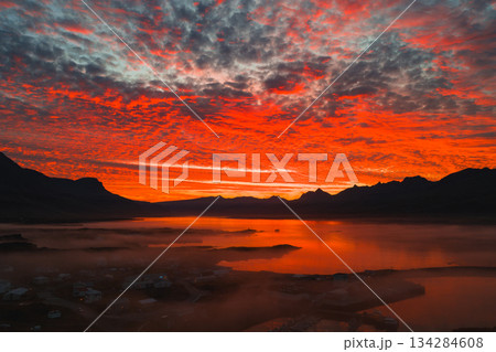 A vivid sunset illuminates the Icelandic sky in red, orange, and pink. Mountains and a calm water body reflect the colors, with mist over a small settlement. A vivid sunset illuminates the Icelandic sky in red, orange, and pink. Mountains and a calm water body reflect the colors, with mist over a small settlement. 134284608
