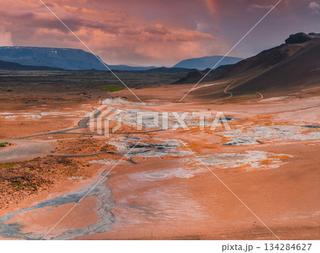 Geothermal area in Iceland with orange and white mineral deposits, steam vents, mud pots, rolling hills, distant mountains, and a winding road. Geothermal area in Iceland with orange and white mineral deposits, steam vents, mud pots, rolling hills, distant mountains, and a winding road. 134284627
