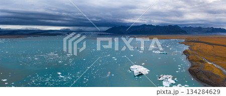 Turquoise waters of Jokulsarlon Glacier Lagoon with floating icebergs, a small boat, a glacier, rugged mountains, and golden brown terrain. 134284629