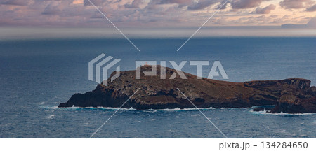A rugged islet with dark cliffs meets white surf at dusk, a small lighthouse atop the highest point near Madeira in Portugal, soft evening light and panoramic view. 134284650