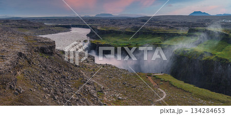 Dettifoss waterfall plunges into a deep canyon with mist rising, surrounded by rocky terrain, green moss, and a pink and blue sky in Iceland. Dettifoss waterfall plunges into a deep canyon with mist rising, surrounded by rocky terrain, green moss, and a pink and blue sky in Iceland. 134284653