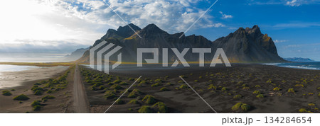 The Stokksnes Peninsula in Iceland features the Vestrahorn mountains, black sand beach, green grass patches, a dirt road, and a partly cloudy sky. 134284654