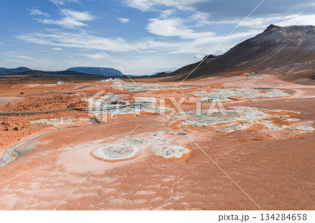 Vibrant geothermal area of Hverir near Lake Myvatn, featuring orange and red earth tones, steaming fumaroles, bubbling mud pots, and a nearby mountain. 134284658