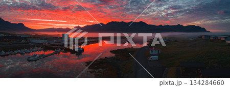 A serene harbor in Iceland at sunset, featuring boats, industrial buildings, calm water, and mist covered jagged mountains in the background. 134284660