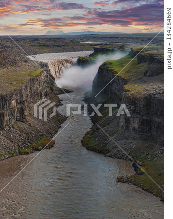 Dettifoss waterfall in Iceland cascades into a rugged canyon, with mist rising and green vegetation on cliffs under a colorful sunset sky. 134284669