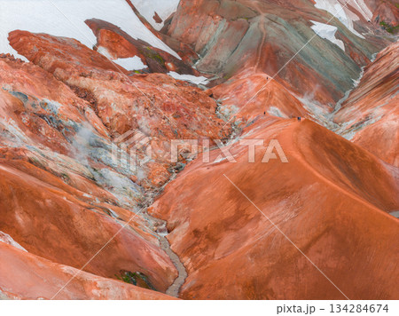 The Rainbow Mountains in Iceland display vibrant red, orange, and green hues from rhyolite rock. Snow patches and a narrow trail highlight the rugged terrain. 134284674