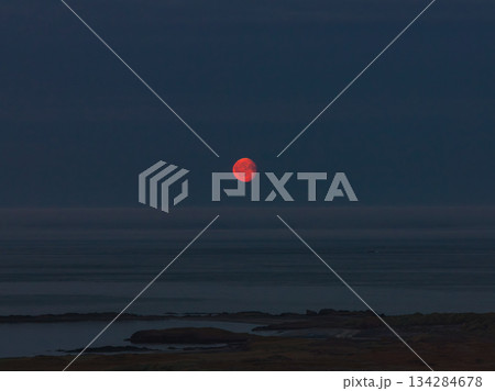 A vivid red moon rises over a calm, dark ocean at twilight, with faint landforms and rocky outcroppings visible along the Icelandic coastline. A vivid red moon rises over a calm, dark ocean at twilight, with faint landforms and rocky outcroppings visible along the Icelandic coastline. 134284678