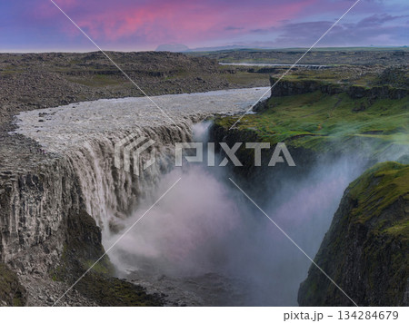 Dettifoss waterfall cascades into a deep canyon with rugged cliffs and green moss, set against a pink and purple sunset sky in Iceland. Dettifoss waterfall cascades into a deep canyon with rugged cliffs and green moss, set against a pink and purple sunset sky in Iceland. 134284679