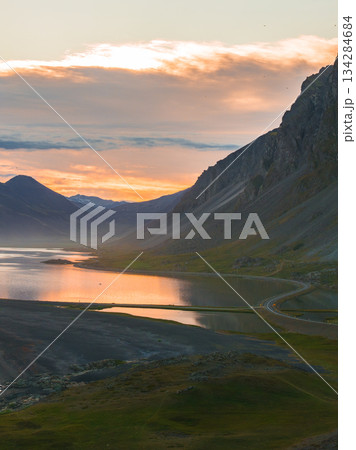 A calm body of water reflects the warm sunset hues, framed by towering mountains. A winding road runs along the right, adding scale to the scene. 134284684