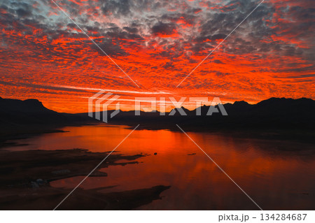 Fiery red and orange sunset reflects on a calm body of water, likely a fjord or lagoon, with dramatic clouds and dark mountain silhouettes in Iceland. Fiery red and orange sunset reflects on a calm body of water, likely a fjord or lagoon, with dramatic clouds and dark mountain silhouettes in Iceland. 134284687