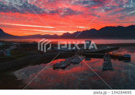 Aerial view of a harbor in Iceland at sunset, featuring docked boats, traditional buildings, calm water, jagged mountains, and a misty horizon. Aerial view of a harbor in Iceland at sunset, featuring docked boats, traditional buildings, calm water, jagged mountains, and a misty horizon. 134284695