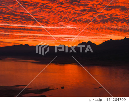 Aerial view of a vivid red and orange sunset reflecting on calm water, with textured clouds and silhouetted mountain peaks in the background, likely in Iceland. 134284709