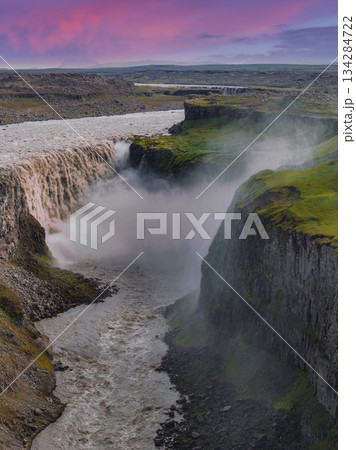Dettifoss waterfall cascades into a deep canyon with moss covered cliffs, mist rising dramatically, under a pink and purple sky in Iceland. 134284722