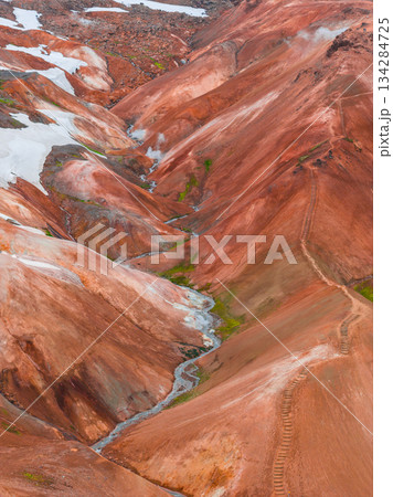 Vibrant red, orange, and brown rhyolite mountains in Landmannalaugar, Iceland, with snow patches, steam vents, a winding trail, and a small stream. Vibrant red, orange, and brown rhyolite mountains in Landmannalaugar, Iceland, with snow patches, steam vents, a winding trail, and a small stream. 134284725