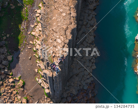 Aerial view of Studlagil Canyon in Iceland, featuring geometric basalt columns, a turquoise river, green vegetation, and people seated on the edge. 134284735