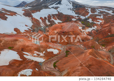Vibrant orange, red, and brown rhyolite mountains with snow patches and a winding trail in Landmannalaugar, Iceland, showcasing unique terrain. 134284740