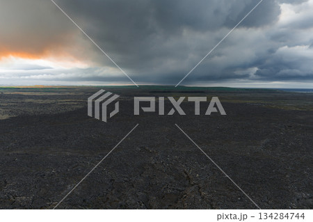 Dark volcanic terrain with cracked surfaces stretches to the horizon under dense clouds. A faint sunlight glow adds contrast to the moody landscape. 134284744