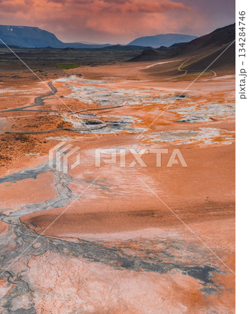 The Hverir geothermal area near Lake Myvatn in Iceland features orange and red terrain, steaming fumaroles, mud pots, and a winding path under a vivid sky. 134284746