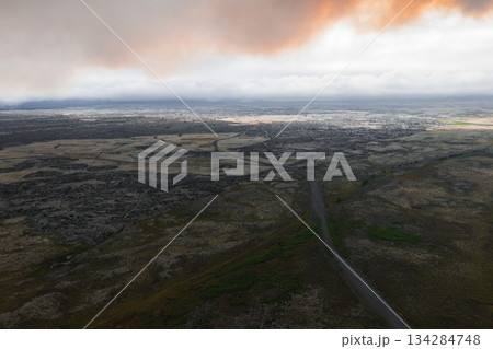 A winding road traverses a barren, rocky terrain with moss patches and sparse vegetation under a dramatic sky with thick clouds and orange glow. 134284748
