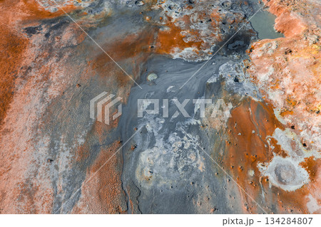 Aerial view of a geothermal area in Iceland featuring vibrant orange, red, and gray hues, steaming vents, bubbling mud pots, and mineral deposits. 134284807
