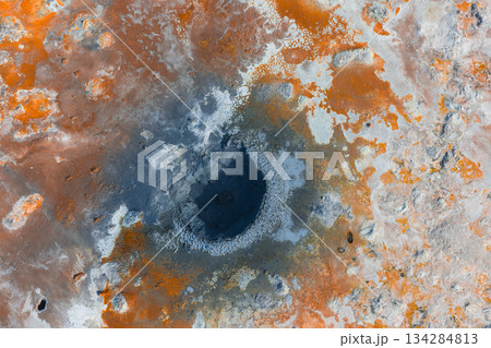Aerial image of a geothermal area in Iceland featuring a dark crater like center surrounded by orange, white, and gray mineral deposits. 134284813