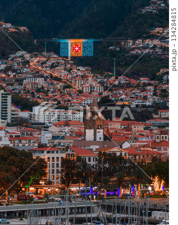 Evening view of Funchal, Madeira, with Se Cathedral centered, terraced red roofs on hillsides, festive lights in flag colors and red cross, marina and promenade aglow. 134284815