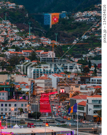 Elevated view of Funchal, Madeira at dusk, red festival lights line an avenue to the harbor, Madeira flag with Order of Christ cross spans slopes, traffic and crowds animate streets. Elevated view of Funchal, Madeira at dusk, red festival lights line an avenue to the harbor, Madeira flag with Order of Christ cross spans slopes, traffic and crowds animate streets. 134284816