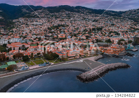 Aerial view of Funchal, Madeira, in evening light, white roofs leading to Praia do Almirante Reis, curved jetty, and Sao Tiago Fortress by the pebble shore. Aerial view of Funchal, Madeira, in evening light, white roofs leading to Praia do Almirante Reis, curved jetty, and Sao Tiago Fortress by the pebble shore. 134284822