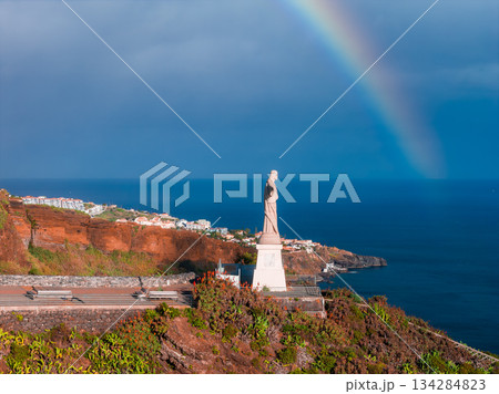 Cristo Rei at Garajau stands on a pedestal above the Atlantic in Madeira, Portugal. Terraced roads, coastal scrub, and white houses appear as a rainbow arcs under post storm light. 134284823