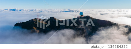 Aerial view shows Pico do Arieiro in Madeira, Portugal, with a blue geodesic radar dome and antennas on a jagged ridge above clouds in cool morning light. Aerial view shows Pico do Arieiro in Madeira, Portugal, with a blue geodesic radar dome and antennas on a jagged ridge above clouds in cool morning light. 134284832