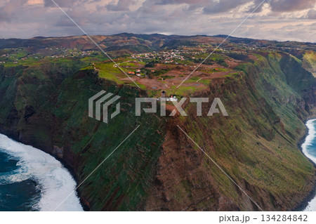 Aerial view shows basalt sea cliffs, terraced fields, and a white lighthouse at Ponta do Pargo, Madeira, Portugal. Late day light and clouds add a moody feel. 134284842
