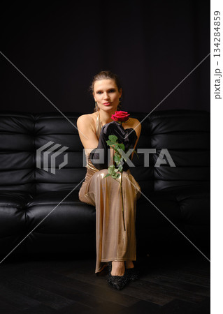Woman holding a red rose while seated on a black sofa during a simple indoor event Woman holding a red rose while seated on a black sofa during a simple indoor event 134284859