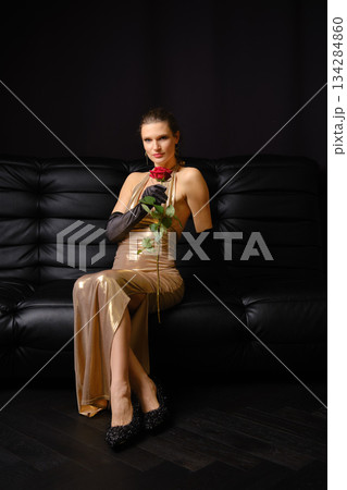Elegant woman holding a rose while sitting on a black sofa at night in a stylish indoor space Elegant woman holding a rose while sitting on a black sofa at night in a stylish indoor space 134284860