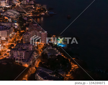 Aerial night scene in Funchal, Madeira, Portugal, shows hotel towers, curving orange streets, and a turquoise circular pool by the Atlantic, with reflections along the coast. 134284871