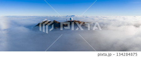 Aerial view shows Pico do Arieiro in Madeira, Portugal, with a white geodesic radar dome and antennas above a cloud layer, rocky ridge in clear daylight. 134284875