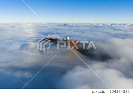 Aerial view shows Pico do Arieiro on Madeira Island with a white radar dome and ridge facilities above clouds at sunrise, soft light on rugged red brown rock and billowing clouds. Aerial view shows Pico do Arieiro on Madeira Island with a white radar dome and ridge facilities above clouds at sunrise, soft light on rugged red brown rock and billowing clouds. 134284882