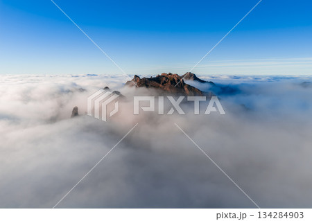 Aerial view shows Pico do Arieiro ridge slicing cloud inversion in Madeira, Portugal. Warm sunrise or sunset light bathes jagged peaks, rocky outcrops, and volcanic forms. Aerial view shows Pico do Arieiro ridge slicing cloud inversion in Madeira, Portugal. Warm sunrise or sunset light bathes jagged peaks, rocky outcrops, and volcanic forms. 134284903
