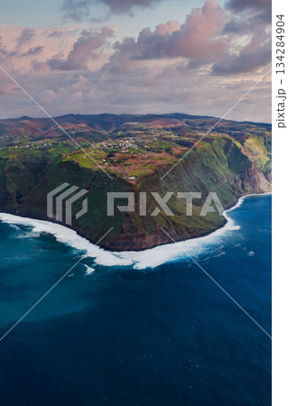 Aerial view of a rugged headland on Madeira Island, Portugal. Sheer green cliffs meet foamy waves, small settlements and terraces sit above in late afternoon light. 134284904