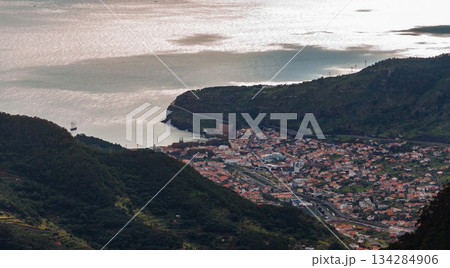 Wide view of a coastal town on Madeira Island, Portugal, terracotta roofs leading to a small harbor and dark promontory, late afternoon light on a calm Atlantic bay. Wide view of a coastal town on Madeira Island, Portugal, terracotta roofs leading to a small harbor and dark promontory, late afternoon light on a calm Atlantic bay. 134284906