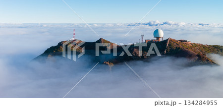 Aerial view shows Pico do Arieiro, Madeira, with white radar dome and towers on a rugged ridge. Warm light hits volcanic peaks as cloud inversion fills the valleys. 134284955