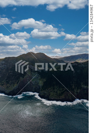 Aerial view of rugged sea cliffs and white surf on Madeira Island, Portugal, dark volcanic shore, green slopes, and high peaks under cumulus clouds in bright daylight. Aerial view of rugged sea cliffs and white surf on Madeira Island, Portugal, dark volcanic shore, green slopes, and high peaks under cumulus clouds in bright daylight. 134284967