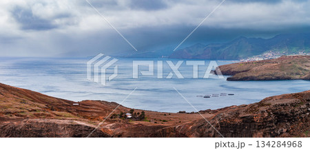 Reddish brown cliffs meet a calm Atlantic bay at Ponta de Sao Lourenco, Madeira, with layered headlands, fish pens, misted mountains, and a distant settlement in overcast light. 134284968