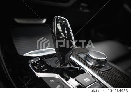 Interior view focusing on a BMW gear selector with labeled automatic transmission modes, glossy black finish, metallic accents, and control buttons. 134284988