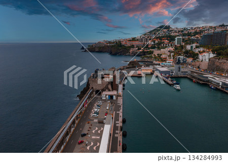 Wide angle view of Funchal harbor, Madeira, Portugal at dusk, with marina boats, fort like stone gateway, terraced houses, hotels, pink clouds, and calm teal water. Wide angle view of Funchal harbor, Madeira, Portugal at dusk, with marina boats, fort like stone gateway, terraced houses, hotels, pink clouds, and calm teal water. 134284993