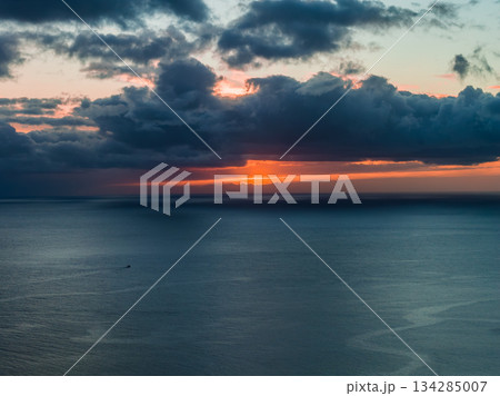Atlantic Ocean at sunset off Madeira, Portugal, with a glowing orange band under dark clouds, subtle ripples, and a tiny boat left, seen from coastal cliffs. 134285007