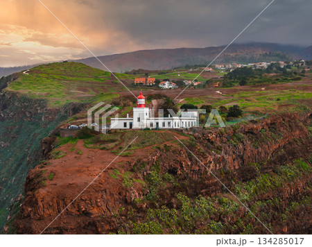 White lighthouse with red lantern on rugged west Madeira cliff, aerial view. Green scrub, scattered buildings, Atlantic beyond, warm low light, wide composition. 134285017