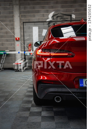 Shiny red BMW X4 parked in a well lit garage with tiled floor, gray brick wall, fire extinguisher, and visible European license plate marked LV. 134285032